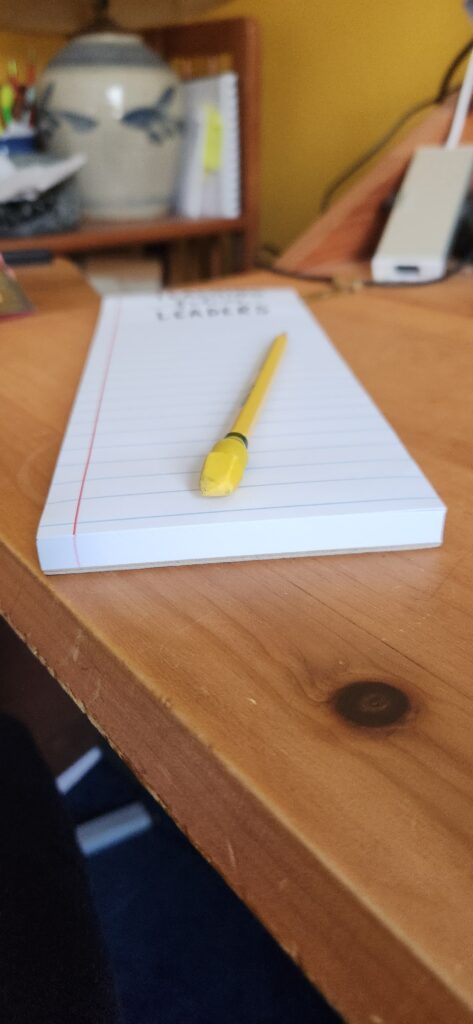 Wooden desk with a long narrow pad resting on it. Resting on the pad is a yellow pencil with a yellow eraser.